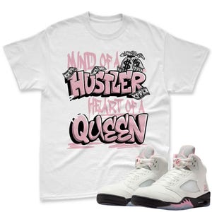 May include: White t-shirt with the text "MIND OF A HUSTLER HEART OF A QUEEN" in pink and black lettering, with dollar bill graphics. The shirt is paired with white and black sneakers with pink accents.