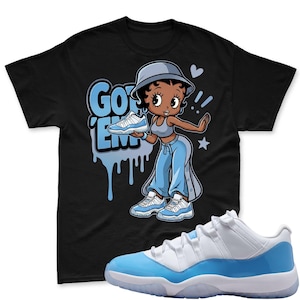 Op de afbeelding: Zwart T-shirt met een cartoonafbeelding van een vrouw die een blauw-witte sneaker vasthoudt, met de woorden "GOT 'EM" in het blauw. Een paar blauw-witte sneakers staan op de voorgrond.