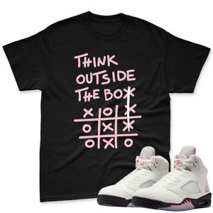 Może przedstawiać: Czarny t-shirt z różowym napisem "THINK OUTSIDE THE BOX" i siatką do gry w kółko i krzyżyk. Białe i różowe trampki z czarnymi podeszwami na pierwszym planie. Trampki mają różowy akcent na podeszwie i języku.