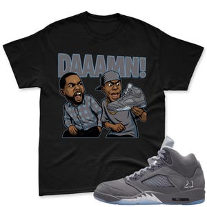 May include: Black t-shirt featuring a cartoon graphic of two men reacting to a gray sneaker. The word "DAAAMN!" is printed above the cartoon. A gray and white Air Jordan 5 sneaker is in the foreground.