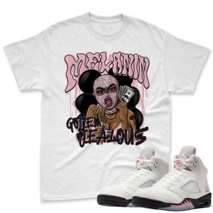 May include: White t-shirt featuring a graphic of a woman in a pink ski mask holding money, with the text "MELANIN" and "GOT EM JEALOUS" in pink and black. White and pink high-top sneakers with black soles are also in the image.