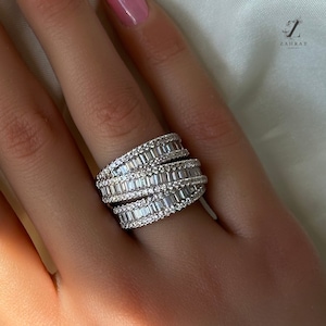 May include: A silver ring with multiple rows of baguette-shaped diamonds. The ring is worn on a person's finger.