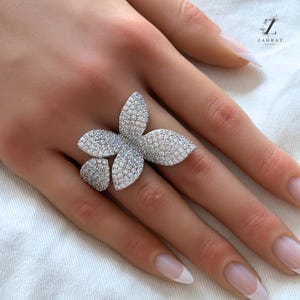 May include: A silver ring featuring a large, flower-shaped design. The ring is set with clear cubic zirconia stones.