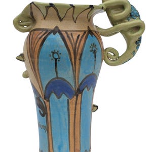 May include: A hand-painted ceramic vase with a unique design. The vase features a light tan base with blue, brown, and green accents. A green chameleon-like figure serves as a handle. The vase has a blue and brown floral pattern and the word "ium" is visible.