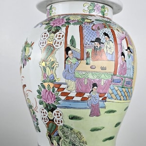 May include: A large, ornate ceramic vase with a lid, featuring a detailed hand-painted scene of figures in a garden setting. The vase is predominantly white with colorful floral and landscape designs, and a gold-colored animal figure on the lid.