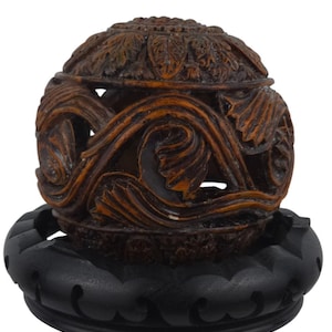 May include: A decorative, carved wooden container with a lid, resting on a black base. The container is brown with intricate swirling patterns and floral details. The base is a solid, dark color.
