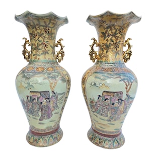 Puede incluir: Par de jarrones antiguos y ornamentados, con una combinación de colores crema y dorado. Cada jarrón presenta una parte superior acampanada, asas doradas y una escena pintada detallada de figuras con vestimenta tradicional. La base tiene un patrón decorativo.