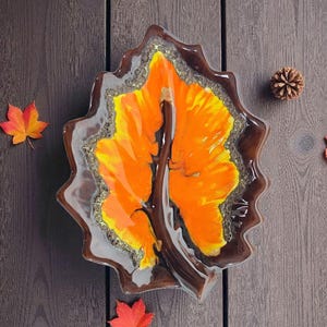 Könnte beinhalten: Eine dekorative Blattschale aus Glas mit einem leuchtenden orange-gelben Design, umrahmt von einem dunkelbraunen Rand. Die Schale ist von Herbstblättern und einem Tannenzapfen auf einer Holzoberfläche umgeben.