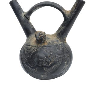 Puede incluir: Un recipiente de cerámica gris oscuro con un cuerpo redondeado, dos picos y un asa. El recipiente presenta diseños en relieve tallados y una pequeña figura con forma de pájaro en la parte superior. La superficie tiene un aspecto envejecido.