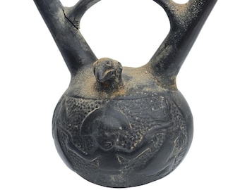 May include: A dark gray ceramic vessel with a rounded body, two spouts, and a handle. The vessel features carved relief designs and a small bird-like figure on top. The surface has a weathered appearance.