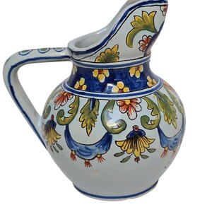 May include: A hand-painted ceramic pitcher with a curved spout and handle. The pitcher features a light blue base with floral and leaf designs in yellow, red, and blue. A dark blue band encircles the top.