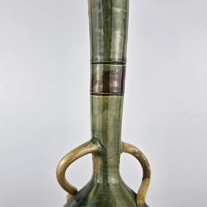 Könnte beinhalten: Eine hohe, grüne Keramikvase mit ausgestelltem Oberteil und zwei gebogenen Griffen. Die Vase hat einen bauchigen Boden und einen langen, zylindrischen Hals mit vertikalen Streifen. Die Vase ist etwa 38 cm hoch.