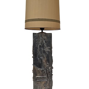 Puede incluir: Una lámpara de pie vintage con una base texturizada de bronce oscuro con tallas en relieve. La pantalla es de color beige claro con un ribete decorativo. La lámpara está diseñada para uso en interiores y proporciona iluminación ambiental.