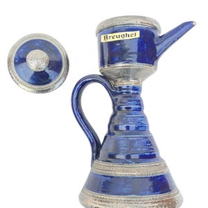 Peut inclure: Une cruche en céramique bleue avec un bec et une anse, accompagnée d'un couvercle. La cruche a un bord argenté et le mot "Breughel" est imprimé sur le côté. Le design est un mélange de bleu et d'argent.