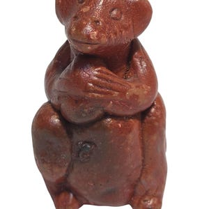 May include: A small, reddish-brown monkey figurine with a seated posture, hands clasped together, and a detailed facial expression. The sculpture has a textured surface and is set against a white background. The figurine is a decorative item.