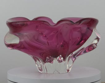Puede incluir: Un cuenco decorativo de cristal con un vibrante tono magenta. El cuenco tiene una forma única y orgánica con un borde ondulado y una base transparente esculpida. El cristal parece tener burbujas de aire.