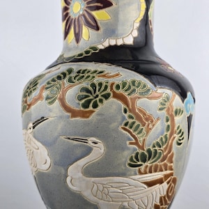 May include: A tall, decorative ceramic vase with a black background and detailed artwork. The vase features white cranes, trees, and floral designs in shades of blue, brown, yellow, and gray. The vase has a blue rim and base.