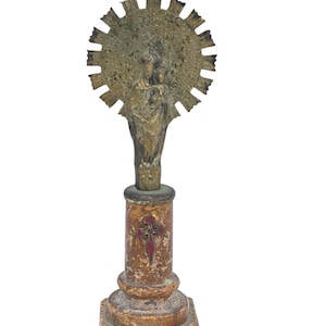 May include: A vintage religious statue depicting a figure holding a child, with a sunburst halo. The statue is mounted on a column with a red cross design, and a square base. The statue is made of a bronze-coloured material.