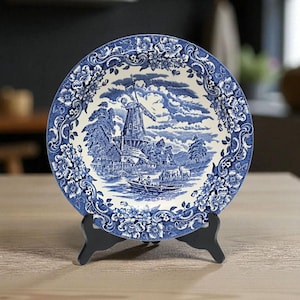 Puede incluir: Un plato decorativo azul y blanco con una escena de molino de viento, exhibido en un soporte negro. El plato tiene un borde floral y una ilustración detallada del paisaje. Es un artículo decorativo.