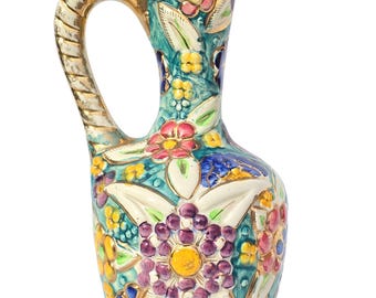 May include: A colorful ceramic pitcher with a gold-trimmed handle and spout. The pitcher is decorated with floral patterns in shades of pink, yellow, blue, and purple against a teal background. The handle is white with gold accents.