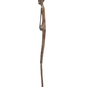 Op de afbeelding: Een gebeeldhouwde houten sculptuur van een slanke menselijke figuur. De figuur heeft een langwerpig hoofd, een smalle torso met een open ruimte en lange, dunne benen. De sculptuur heeft een natuurlijke bruine kleur en staat op een kleine, rechthoekige basis.