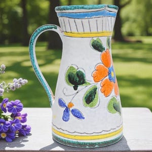 Könnte beinhalten: Ein weißer Keramikkrug mit floralem Design. Der Krug hat orange, blaue und grüne Blumen mit blauen und gelben Bändern um den oberen und unteren Rand. Der Griff ist grün und der Krug steht auf einer Holzoberfläche.