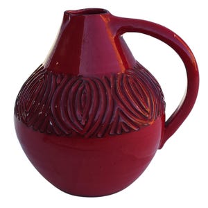 May include: A glossy, deep red ceramic pitcher with a rounded body and a curved handle. The pitcher features a textured, carved design around the middle. The spout is small and the handle is large enough to grip comfortably. The pitcher is isolated on a white background.