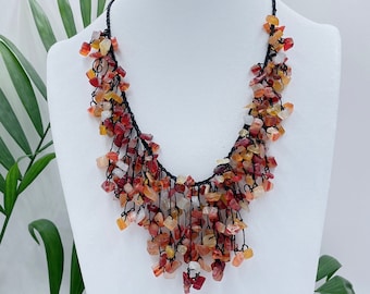 May include: A red, orange, and white gemstone necklace with a black cord. The necklace is made of small, irregular shaped stones that hang from the cord.