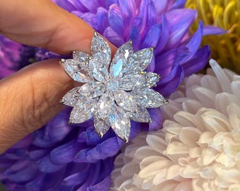 Könnte beinhalten: Ein silberner Ring mit einem großen, blütenförmigen Design. Der Ring ist mit zahlreichen klaren, funkelnden Edelsteinen besetzt, die wie eine blühende Blume angeordnet sind. Der Ring befindet sich vor einem Hintergrund aus lila, weißen und gelben Blumen.