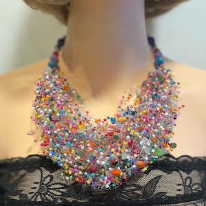 Puede incluir: Un collar de cuentas colorido con un diseño amplio y en cascada. El collar presenta una variedad de pequeñas cuentas redondas en tonos rosa, azul, amarillo, verde y naranja, intercaladas con piedras más grandes de forma irregular. Las cuentas están ensartadas con un alambre plateado.