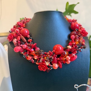 May include: A striking red beaded necklace on a black display stand. The necklace is composed of various red beads, including large spherical beads, small seed beads, and rose-shaped beads, arranged in a layered pattern. The necklace is a statement piece.
