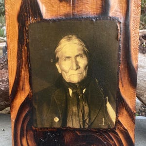 May include: A framed black and white photograph of an older man with long white hair and a serious expression. The frame is made of wood with a dark brown stain and a rough texture. The photo is mounted on a dark brown background.