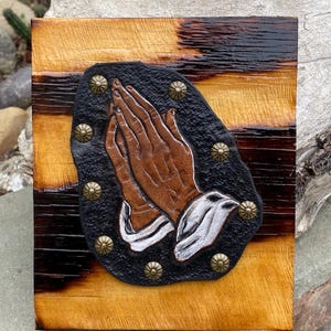 May include: A wooden plaque with a black leather background and a brown leather image of two hands clasped in prayer. The hands are surrounded by decorative metal studs.