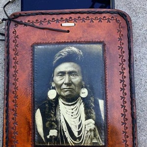 May include: A brown leather case with a picture of a Native American man wearing a beaded necklace and earrings. The case has a decorative border and a flap that closes with a button.
