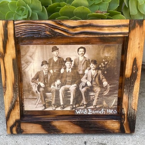 May include: A rustic wooden frame with a black and white photo of five men in suits and hats. The photo is titled "Wild Bunch 1900".