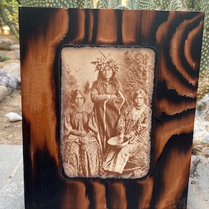 May include: A framed sepia-toned photograph of three Native American people. The person in the center is wearing a feathered headdress and has their arms crossed. The people on either side are wearing traditional clothing and jewelry.