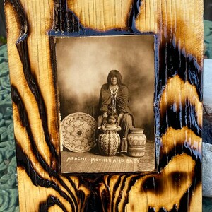 May include: A framed black and white photograph of an Apache mother and baby. The mother is wearing traditional clothing and holding a baby in her arms. The photo is set against a wooden background with a black border.