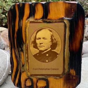 May include: A framed black and white portrait of Col. Christopher Carson in a military uniform. The frame is made of wood with a burnt finish.