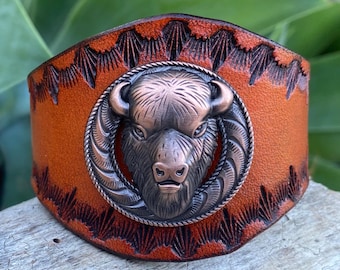 May include: A wide, brown leather cuff bracelet with a detailed, copper-colored buffalo head emblem. The leather is embossed with a decorative pattern around the edges. The cuff is designed to be worn on the wrist.