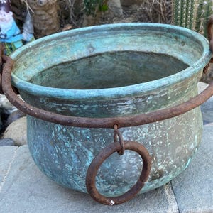 May include: A weathered copper cauldron with a rounded body and a wide, open top. The exterior has a blue-green patina, and a rusty metal handle arches over the opening. A smaller ring hangs from the center of the handle. The cauldron sits on a stone surface.