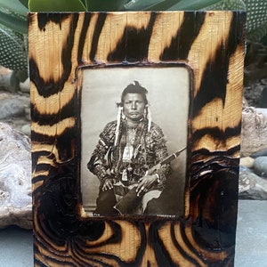 May include: A black and white photograph of a Native American man wearing traditional clothing and holding a rifle. The photo is framed in a wooden frame with a burnt wood finish.