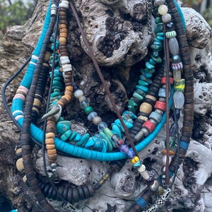 May include: A collection of layered necklaces with various beads and materials. Turquoise, wooden, and seed bead necklaces are visible, along with a few with metal accents. The necklaces are displayed on a piece of driftwood.