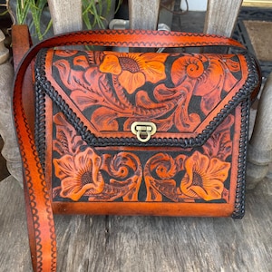 May include: A handcrafted leather crossbody bag with a floral design. The bag features a flap closure with a metal clasp, a braided black border, and a long adjustable strap. The embossed flowers are orange against a dark brown background.
