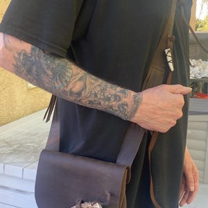 May include: A brown leather shoulder bag with a flap closure and fringe detailing. The bag features a decorative antler piece on the front and a long strap. The bag is worn over a black shirt.
