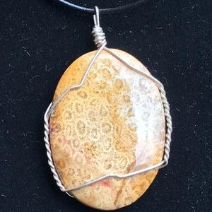May include: Oval pendant necklace featuring a polished, patterned stone in shades of tan and cream, encased in a silver wire frame. The pendant hangs from a black cord, creating a simple, elegant design. The stone's intricate pattern is visible.