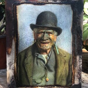 May include: A framed portrait of a man wearing a black bowler hat and a green jacket. He has a traditional Maori tattoo on his face and a green and blue feather earring.