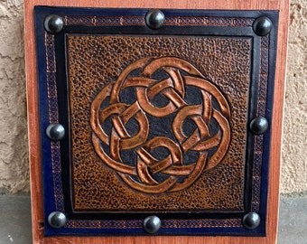 May include: A square wooden plaque with a dark blue and black border. The plaque features a copper-colored Celtic knot design in the center.