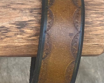 May include: A brown leather bracelet with a black edge and a decorative pattern of circles.