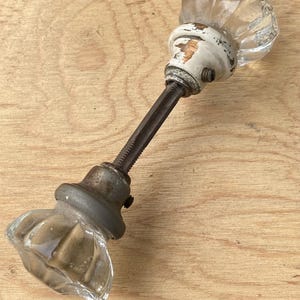 May include: Antique glass door knob with a clear, ribbed design. The knob has a white and aged metal base and a long, threaded rod. The hardware shows signs of wear, adding to its vintage appeal. Perfect for adding a touch of nostalgia to any door.