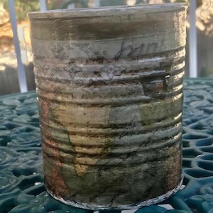 May include: A repurposed tin can with a decorative design. The can is painted with a multi-colored pattern and has a distressed look. The can is sitting on a blue and green surface.